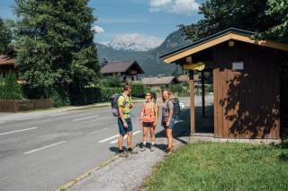Drei Wander*innen an einer Bushaltestelle vor Bergpanorama