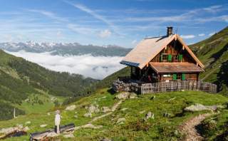 Eine holzvertäfelte Spitzdach-Hütte steht auf einer Anhöhe über einem nebelverhangenen Tal, darüber ein sonniger Tag.