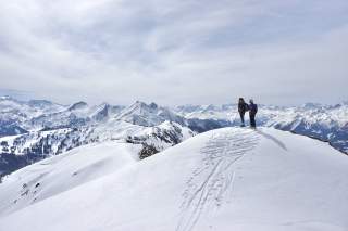 Zwei Menschen mit Tourenski stehen auf schneebedeckter Kuppe mit Ausblick auf verschneite Gipfel