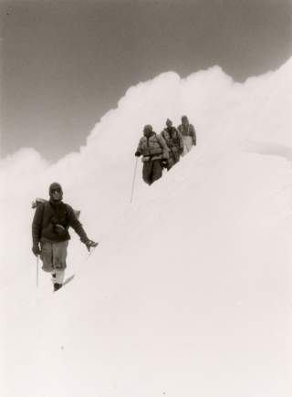Vier Menschen in steilem verschneitem Gelände, Schnee und Wolken gehen ineinander über.