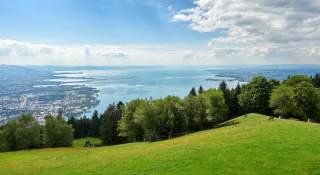 Blick von einer Bergwiese auf einen großen See, links im Tal eine Stadt, im Hintergrund kleine Berge.