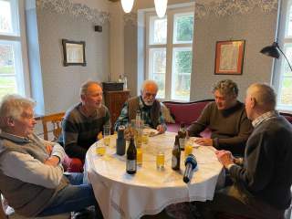 5 Männer sitzen an einem runden Tisch mit einer weißen Tischdecke, auf dem Tisch steht Orangensaft und Bier.