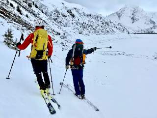 Zwei Menschen auf Tourenski, einer zeigt mit Skistock Richtung Staumauer eines Sees