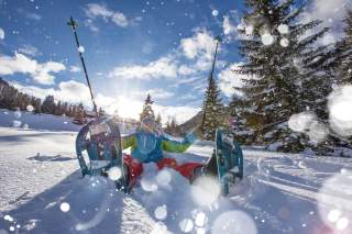 Mensch mit Schneeschuhen sitzt im Schnee und streckt Stöcke in die Höhe