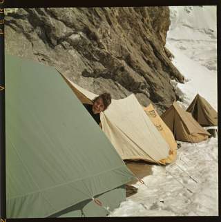 einige Zelte sind an einem Felsvorsprung zwischen Wand und Schnee aufgebaut. Eine Frau streckt den Kopf aus einem Zelt und lacht.