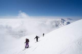 Drei Skitourengeher queren einen Hang mit windgepresstem Schnee, Im Hintergrund durchbrechen Berggipfel die Wolkendecke.