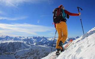 Frau geht auf Tourenski steilen Schneehang hinauf mit Blick auf verschneite Alpenkette