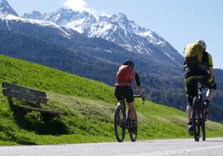 Zwei Radfahrer fahren aus dem Bild, im Hintergrund stehen Berge mit schneebedeckten Gipfeln